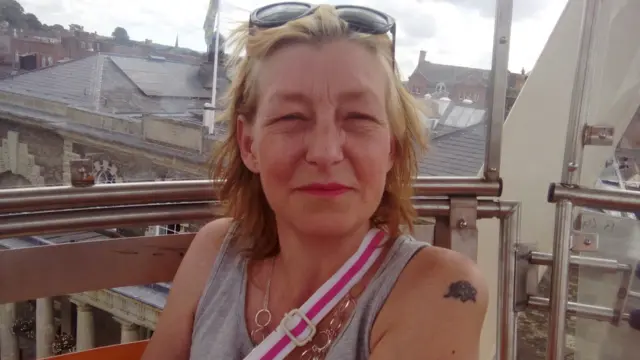 Dawn Sturgess in a grey vest with a pink and white bag over her shoulder, sitting near a window.