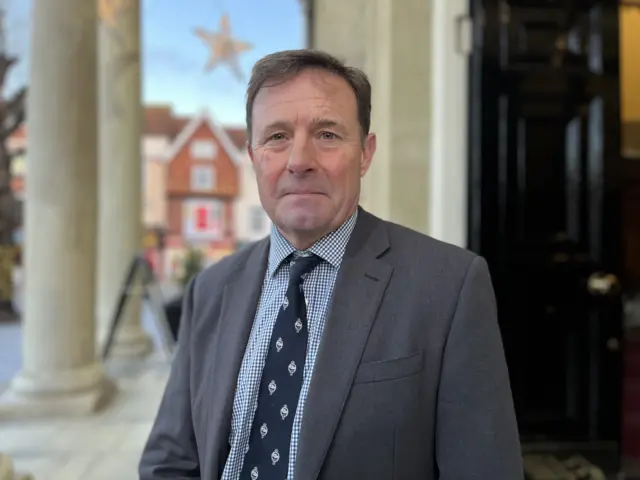 Hamish De Bretton-Gordon wearing a grey blazer with a blue and white checked shirt and navy tie. He is standing outside a building with large pillars showing behind him.