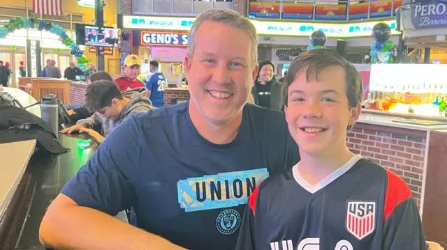 A man and his son smile while wearing blue football shirts