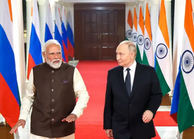 Putin on the right, see in a black suit and navy tied, walking on the red carpet with Modi who is wearing a cream kurta with a brown jacket over it. The two leaders are walking along a corridor lined with Indian and Russian flags