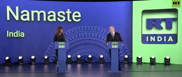 Editor-in-chief Margarita Simonyan and Putin on stage behind blue podiums. The screen behind them says Namaste India and features the RT India logo in blue and green