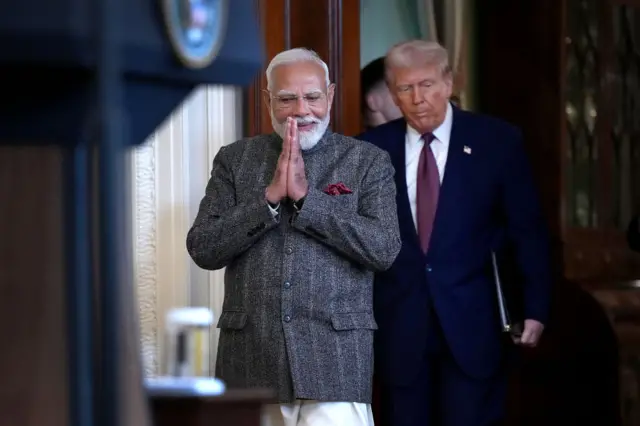 Modi with his hands folded, wearing a grey striped jacket as Trump walks behind him in a navy suit, holding a folder in his left hand