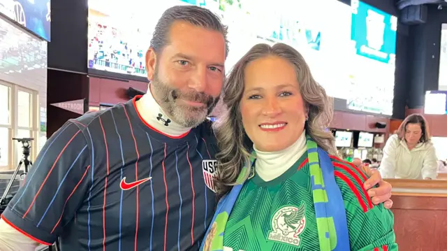 A man in a USMNT soccer shirt and a woman in a green Mexican national team shirt with a scarf