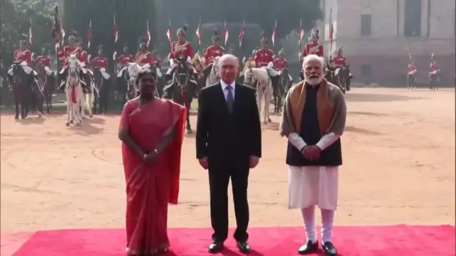 President Murmu on the left in a red sari next to Putin in black suit and blue tie with Modi on the right in a white kurta, black jacket and brown and grey shawl. They are standing on a red carpet before the guard of honour