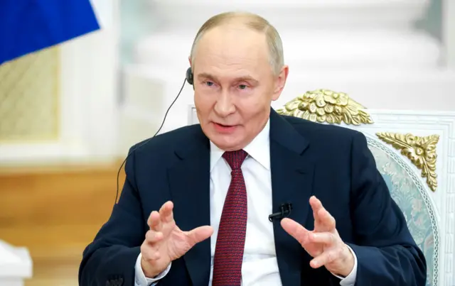 Putin seen wearing an earpiece as he talks. He's wearing a navy blue suit with checked red tie and white shirt