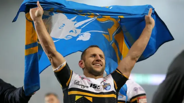 A rugby player lifts a blue and yellow flag with a white Rhino on it above his head