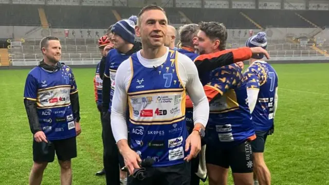 A man stands in a stadium on the pitch in the rain. A group of people behind him, some of them hugging.