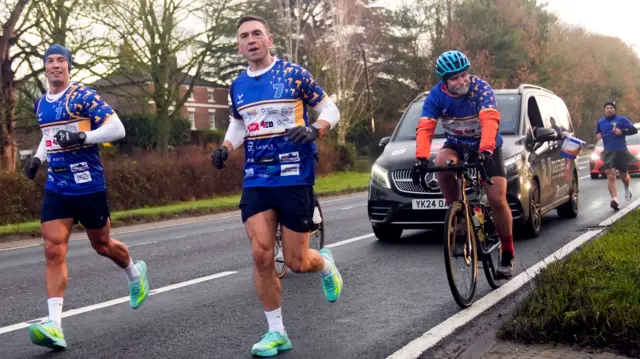 Three men running in blue tops and shorts. Two cyclists on the road behind them and two cars. One car has a person holding a fundraising bucket outside the window.