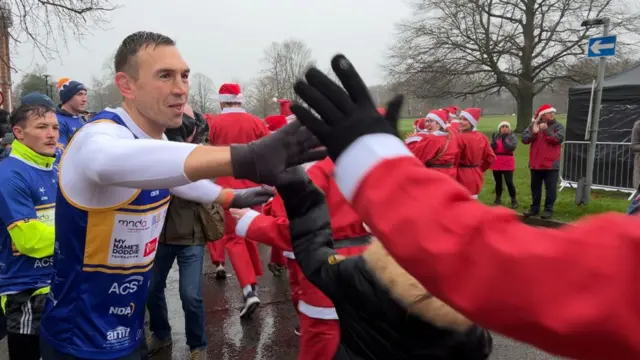 A man in running clothes high fives a man dressed as Santa. A crowd behind him, many dressed in Santa outfits.