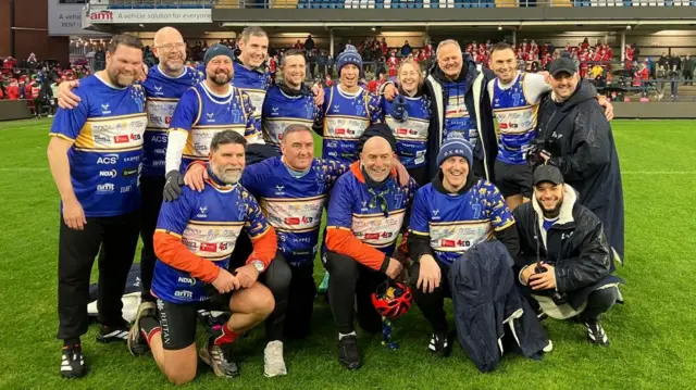 14 men pose together on a rugby pitch wearing blue jerseys