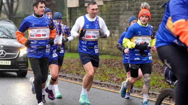Three men running side by side on a road in the rain. A black car behind them.