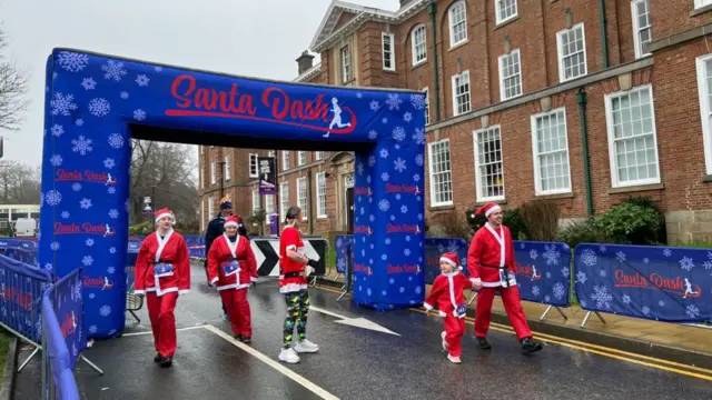 A large blue banner across a road with the sign saying Santa Dash. Five people dressed as Santas are walking underneath the arch.