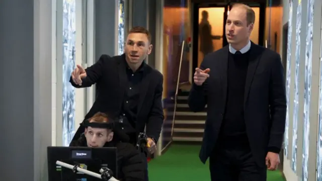 Three men in the corridor of a sports stadium. Sinfield and Prince William stand pointing towards the camera, while Rob Burrow is sitting in his wheelchair.