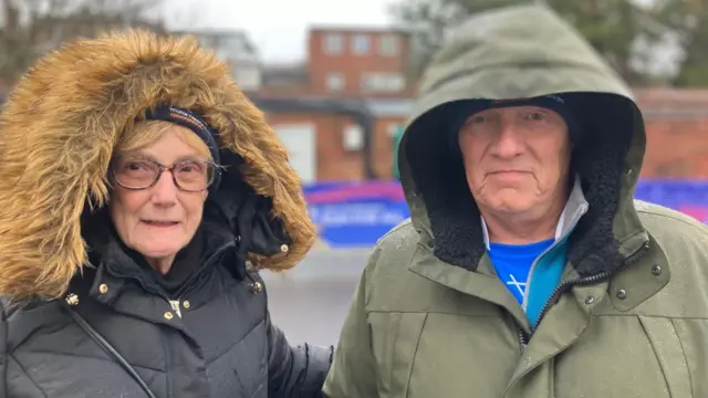 Two people standing outdoors on a wet day, dressed in heavy winter coats with hoods up. One coat is black with a fur-lined hood, and the other is green with a plain hood.