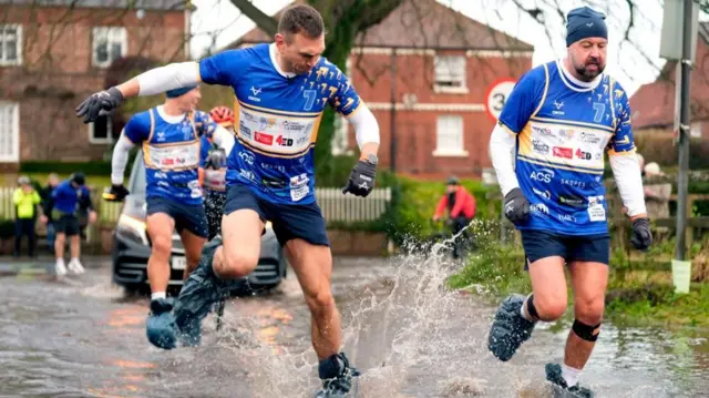 Kevin Sinfield and another man run through puddles in blue, white and yellow sports kits and bin bags over their feet.