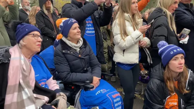 A group of people gathered outdoors, some wearing blue and orange clothing and accessories associated with motor neurone disease awareness. Two individuals in the foreground are seated in wheelchairs, covered with blue blankets.