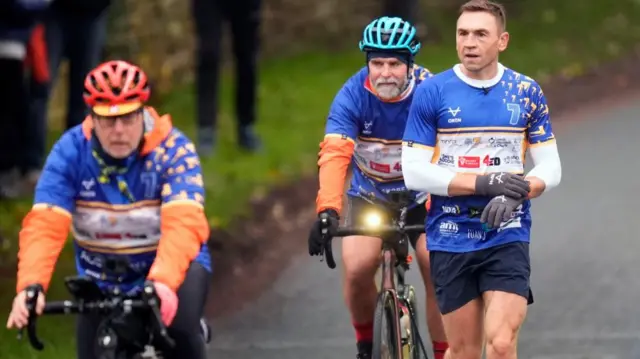 Two men on a bike and one running. They are wearing blue, orange and white sports gear.