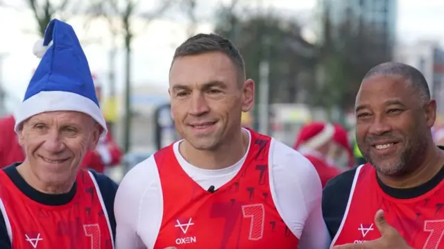 Three men smile at the camera wearing red vests. One of them has a blue Santa hat