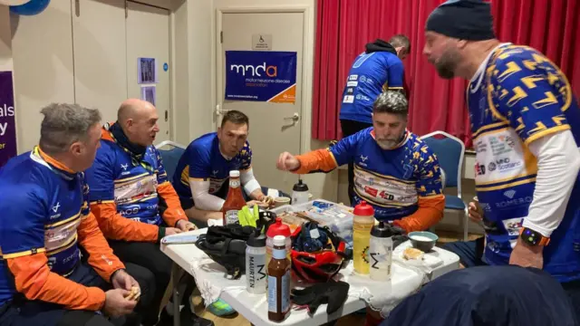 Five men sit around a white table eating and drinking. They are all wearing sports gear