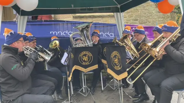 A brass band wearing black jackets and trousers sitting under a marquee