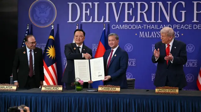 The leaders of Thailand and Cambodia stand side-by-side, shaking hands and holding a document. Trump claps his hands as he watches from the side.