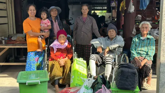 A family with two elderly people in wheelchairs looking at the camera as they wait in front of their house.