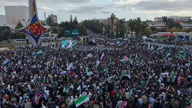 People with flags, including Syrian flags, gather in front of a screen in Umayyad Square in Damascus