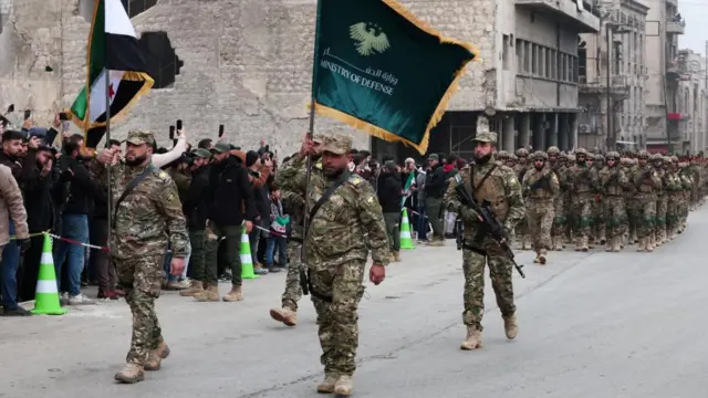 Members of the military, some carrying guns, others carrying flags, file past civilians during a military parade on the streets of Aleppo