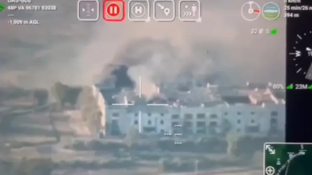 A frame grabbed from the video that shows information around the screen and cross hairs targeting a white building in the middle. Smoke can be seen issuing from the building in the footage