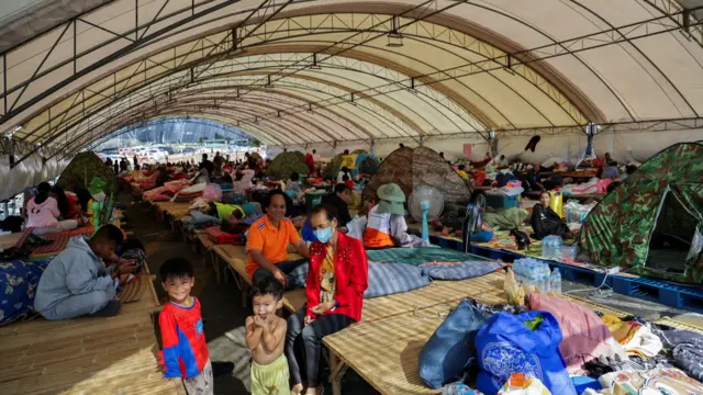 Lots of people in a shelter that has an arched roof. there are tents set up inside it and mats placed on the ground