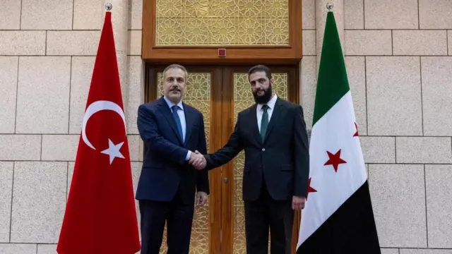 Fidan and al-Sharaa shake hands in front of a Turkish flag and a Syrian flag. Both men wear suits, shirts and ties