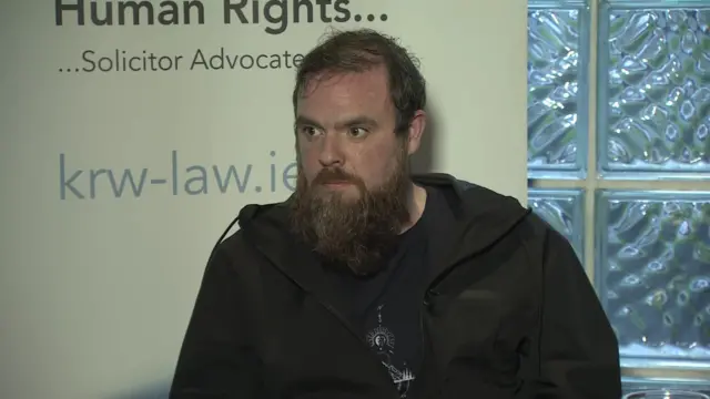 A Man is seated with a long brown beard and short brown hair. He is wearing a black windbreaker with a graphic black tee underneath.