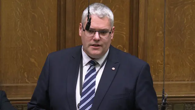 Gavin Robinson standing in the Commons. He is wearing glasses, a suit and striped navy tie.