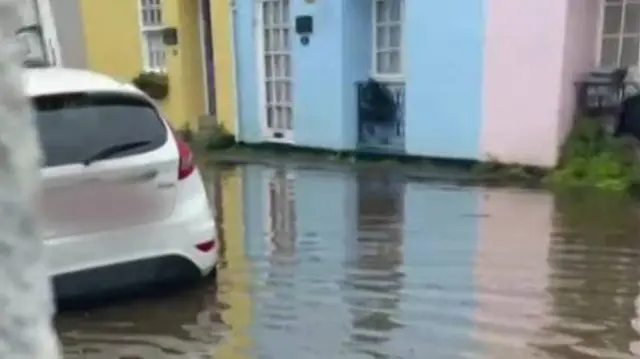 A street full of flood water, up to around the middle of a car's wheel that is parked on the road