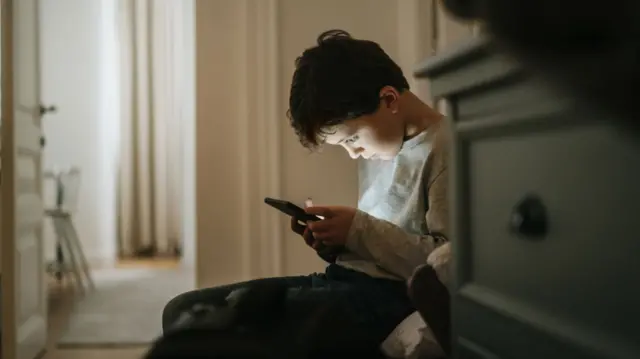 A boy stares at his phone screen while sat at home