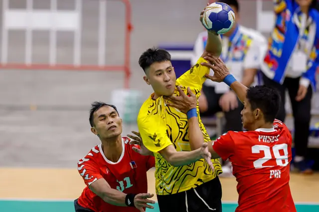 An athlete in a yellow shirt holds a handball aloft while being pushed from either side by two athletes in red shirts