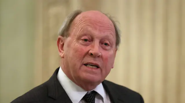 Allister is partially bald with some hair and the side and is speaking in the pic. He is wearing black blazer, pattern tie and white shirt.