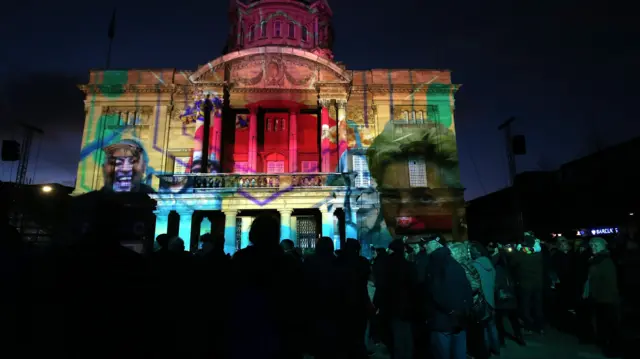 Some illuminations projected onto a town hall at night. Crowds of people, mainly cast in shadow, watch on.