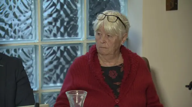Woman sits in press conference. Wearing a red cardigan, she has short blonde hair with glasses atop her head. A glass jug sits in front of her.