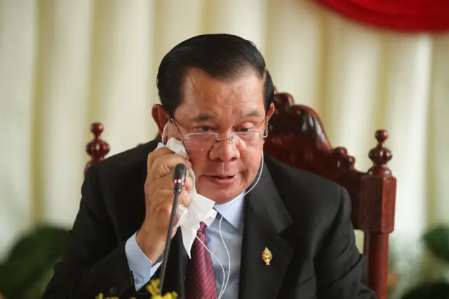 Man in suit and glasses seated and wiping his face with white tissue