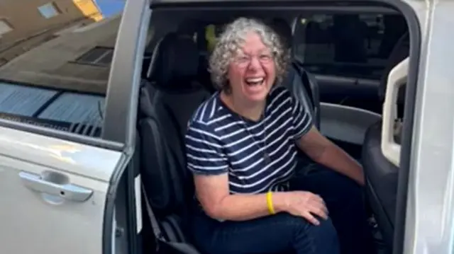 A woman in a stripy top smiling in a car which has the door open.
