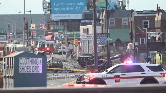 Police car blocking a road, with the crash scene behind it and burnt out cars.