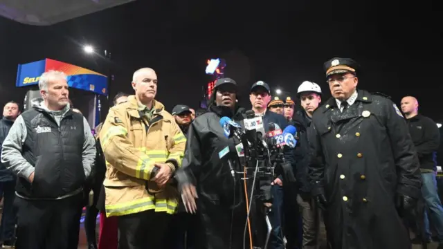 A group of officials gathered around a group of press microphones. The mayor is in the centre, wearing a black cap and black rain jacket, the chief of police to her left and a firefighter in yellow jacket to her right