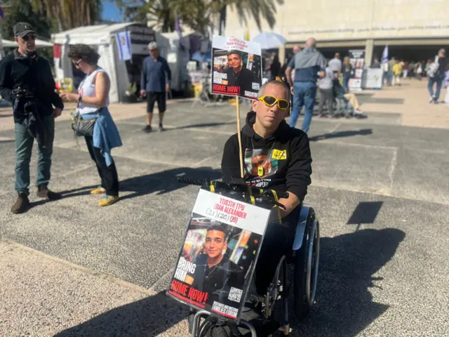 Doron Zexer, in a wheelchair, holds a large photo of Israeli-American hostage Edan Alexander at Hostages Square in Tel Aviv