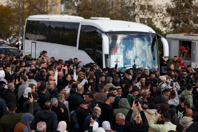 A white bus surrounded by people. It has a red cross symbol in the window