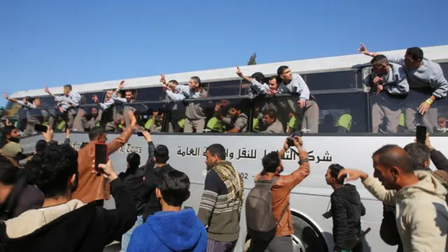 A bus carrying people wearing grey jumpers hang outside the windows waving at a crowd outside.