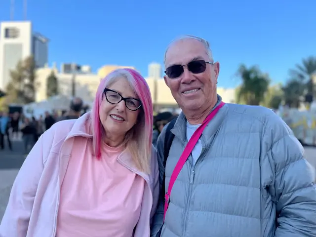 A woman in a pink tshirt and white jumper stands next to man in a grey coat, looking at the camera, with blurred background