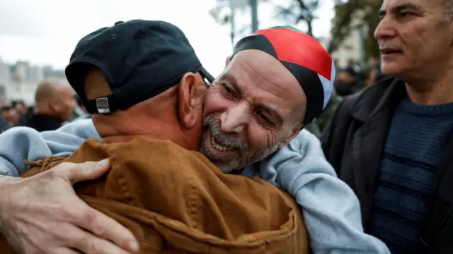 A man in grey hoodie hugs another man with smile on face