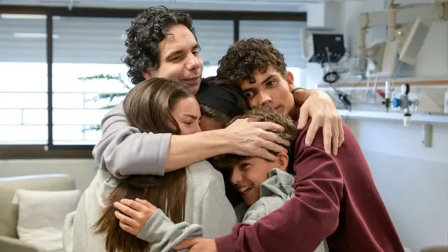 A man hugging four people. One woman's face is covered but you can see the faces of two boys and a girl. One boy smiles, the others look emotional.