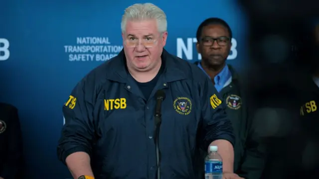 Todd Inman in a jacket with NTSB written on it with hands on a desk talking into a microphone